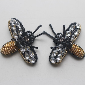 May include: Two black and gold bee-shaped embellishments with clear crystal rhinestones and black beads. The bees have a gold beaded body and black beaded legs and wings.