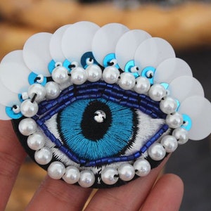 May include: A blue evil eye charm with white pearl beads and white sequins. The charm is embroidered with blue thread and has a black centre.
