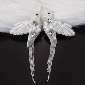 May include: A pair of white hummingbird-shaped earrings. The earrings are embellished with white beads, crystals, and dangling rhinestone chains. The wings are detailed with small white beads. The earrings are set against a white fluffy background.