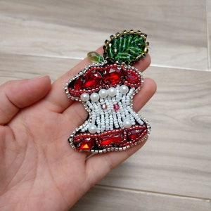 Pode incluir: Uma maçã vermelha e branca de contas com uma folha verde. A maçã é um broche e tem um alfinete na parte de trás.