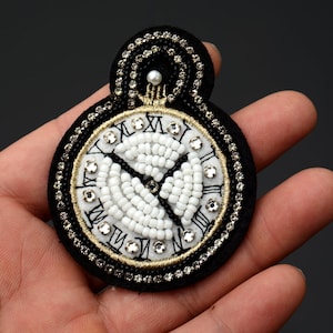 May include: A black and white beaded clock face with Roman numerals and a pearl accent. The clock face is surrounded by a black fabric with rhinestones.