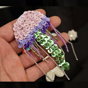 May include: A handmade, sequined jellyfish brooch in shades of pink, purple, and green. The brooch features rows of circular sequins and beaded strands, with a white fabric base. The design is held in a hand against a dark background.