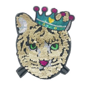 May include: A gold sequined tiger face with a teal blue crown with a yellow heart on top. The crown has pink and green accents.