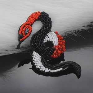 Puede incluir: Broche de serpiente con cuentas en diseño en espiral. La cabeza es roja, con un ojo negro y una lengua negra. El cuerpo es negro, blanco y rojo, con una mezcla de cuentas y lentejuelas. El broche está sobre una superficie reflectante.