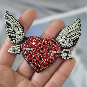 May include: A red heart-shaped patch with rhinestones, surrounded by two wings with rhinestones. The wings are black with silver trim.