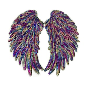 May include: Two iridescent sequined angel wings. The wings are a mix of purple, blue, and gold sequins, arranged to mimic feathers. The wings are symmetrical and set against a white background.