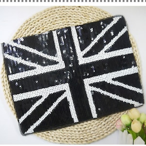 May include: A rectangular clutch with a black sequined surface, featuring a white Union Jack design. The clutch is set against a woven, beige background, with a small floral arrangement in the corner.
