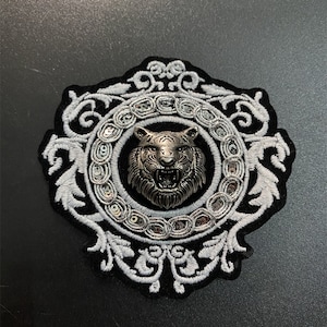 Puede incluir: Un parche decorativo con una detallada cabeza de tigre plateada en el centro, rodeada por un marco circular con lentejuelas plateadas. El marco está sobre un fondo negro con bordados blancos ornamentados. El parche tiene un borde negro.