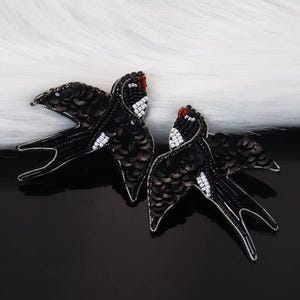 May include: Two black sequined bird brooches with white and red detailing. The birds are made with small sequins and beads, set against a black and white background. These brooches are likely for clothing or accessories.