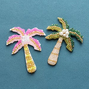 May include: Two beaded palm tree ornaments, one with pink and yellow beads and the other with green and white beads. The palm trees are on a blue background.