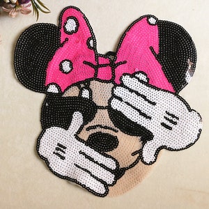 May include: A sequined Minnie Mouse patch featuring a pink bow with white polka dots, black ears, and white gloves covering the eyes. The patch is made with black, white, and pink sequins, creating a sparkling effect. The design is a playful and decorative element.