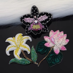May include: Three beaded floral patches: a black orchid with pink and yellow accents, a white and yellow lily, and a pink lotus flower. The patches are intricately detailed with beads and feature green leaves and brown stems.