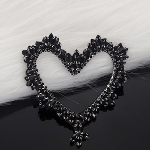May include: A black heart-shaped embellishment, encrusted with dark gemstones. The openwork design features a detailed border of small, sparkling stones. The heart is set against a backdrop of white fur and a glossy black surface.
