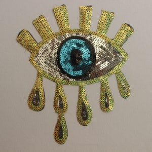 May include: A gold sequin eye with a blue sequin pupil and gold sequin teardrop accents.