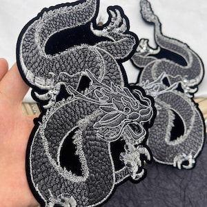 May include: Embroidered dragon patches in black and silver. The patches feature detailed stitching, with the dragon's scales and features highlighted in silver thread. The patches are on a black velvet backing.