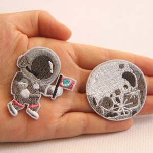 Könnte beinhalten: Gestickte Aufnäher mit einem Astronauten, der eine Flagge hält, und einem detaillierten Mond. Der Astronaut trägt einen grauen Raumanzug mit rosa Akzenten. Der Mond-Aufnäher ist grau und weiß mit Kratern. Ideal für Weltraum-Enthusiasten.