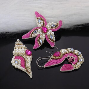 Può includere: Tre applicazioni in tessuto rosa e oro decorate con perle e strass. Le applicazioni hanno la forma di una stella marina, una conchiglia e una coda di sirena.