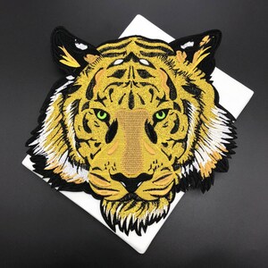 May include: Embroidered tiger head patch with gold, black, and white thread. The tiger has green eyes and a detailed design. The patch is on a white surface.