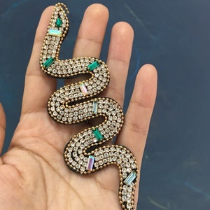 Op de afbeelding: Een gouden broche in de vorm van een slang met heldere en groene strass steentjes. De broche is op een hand.