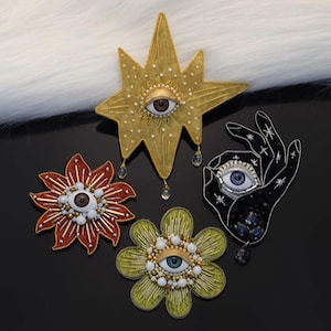 May include: Four ornate brooches with eye designs. A gold star, a red flower, a yellow flower, and a black hand. Each brooch features an eye with detailed iris and embellishments. The star has dangling teardrop accents.