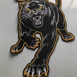 Könnte beinhalten: Gestickter Aufnäher einer schwarzen Panther mit gelben und weißen Akzenten. Die Raubkatze wird in einer knurrenden Pose mit ausgestreckten Krallen dargestellt. Der Aufnäher hat einen gelben Rand und ist für Jacken, Taschen oder andere Kleidungsstücke geeignet.