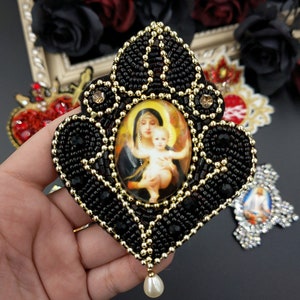 May include: A black beaded and gold-trimmed religious icon with a pearl drop. The icon features a depiction of the Virgin Mary holding the baby Jesus.