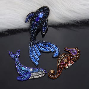 May include: Three beaded aquatic creature embellishments: a blue fish, a blue whale, and a seahorse. The fish has a dark blue body with blue sequins and a large blue gem. The whale is blue with white accents. The seahorse is brown and purple.
