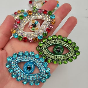 May include: Four colourful evil eye charms with rhinestones and a silver-toned frame. The charms are in shades of blue, green, pink, and orange.