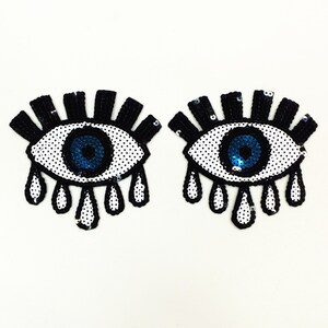 Può includere: Due toppe per occhi nere e bianche con paillettes blu. Ogni occhio ha un design a lacrima con paillettes bianche.