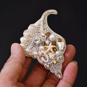 May include: A seashell-shaped brooch featuring intricate beadwork, white pearls, clear faceted crystals, and small brown accents. The design incorporates a miniature starfish and a small cowrie shell, all arranged on a light-colored base.