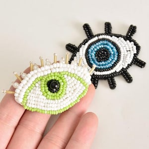 May include: Two beaded evil eye charms. One is white with a green eye and the other is black with a blue eye.