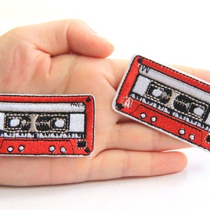 May include: Two embroidered patches in the shape of a retro cassette tape. The patches have a red border and a white and black interior design. The patches are held in a person's hand.