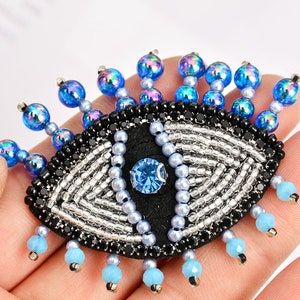 May include: A beaded eye with a blue centre and silver and black beads surrounding it. The eye is surrounded by a fringe of blue and iridescent beads.