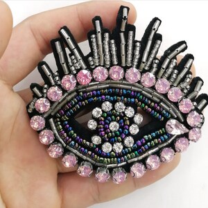 May include: A black, beaded eye patch with pink and clear crystal accents. The eye design features a black velvet base, surrounded by iridescent beads and silver-coloured rods. Pink crystals encircle the eye, with clear crystals in the centre.