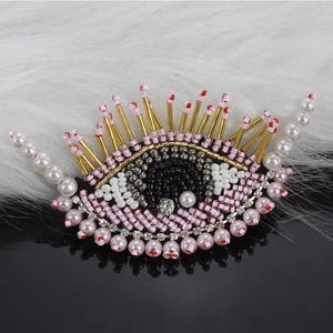 May include: A pink and black beaded eye with gold accents. The eye is surrounded by a ring of white and pink beads.