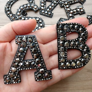 May include: Close-up of the letters A and B, encrusted with dark grey rhinestones. The letters are on a black backing. Other letters are visible in the background, suggesting a set of alphabet letters.