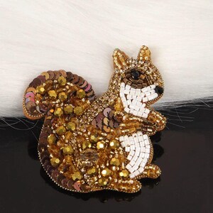 Peut inclure: Une broche en forme d'écureuil réalisée avec un travail complexe de perles et de paillettes. Le motif présente un mélange de perles dorées, brunes et blanches, avec des paillettes brun foncé formant la queue et le corps, et un poitrail blanc.