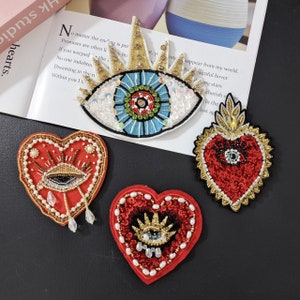 May include: Four embroidered patches featuring an eye motif. One patch is a blue eye with gold rays, and the other three are red heart-shaped patches with gold accents and an eye in the centre.