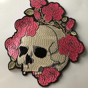 Può includere: Una toppa ricamata raffigurante un teschio umano beige circondato da rose rosa acceso e foglie verdi. Il disegno è delineato con filo nero, mostrando cuciture dettagliate che mettono in risalto i tratti del teschio e la composizione floreale.