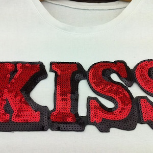 以下が含まれることがあります： 大きな赤いスパンコールで「KISS」の文字が描かれた白いTシャツ。文字は黒いスパンコールで縁取られています。Tシャツはクルーネックで、スパンコールは密に配置され、大胆で人目を引くデザインです。