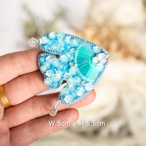 Può includere: Spilla blu fatta a mano con un design a farfalla. La spilla è adornata con piccoli fiori bianchi, perline e un elemento centrale a forma di ventaglio. La spilla misura 5 cm di larghezza e 5,5 cm di altezza.
