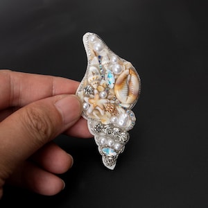 Peut inclure: Broche en forme de coquillage ornée de divers coquillages, perles et cristaux scintillants. La broche est blanche avec une bordure argentée, sur fond noir.