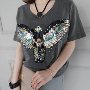 May include: A grey V-neck t-shirt featuring a sequined and beaded design of a stylized bird or insect. The design incorporates black, gold, teal, and white sequins, with small pearl-like beads. A black necklace with a gold pendant is also visible.