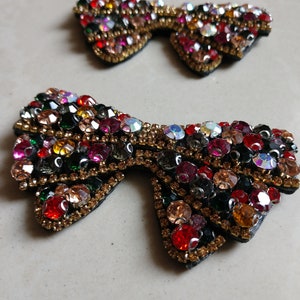 May include: Two bow-shaped embellishments, each decorated with an array of colourful rhinestones. The rhinestones are in shades of red, orange, green, and clear, set against a black base with gold trim. These decorative pieces are ideal for adding sparkle to clothing or accessories.