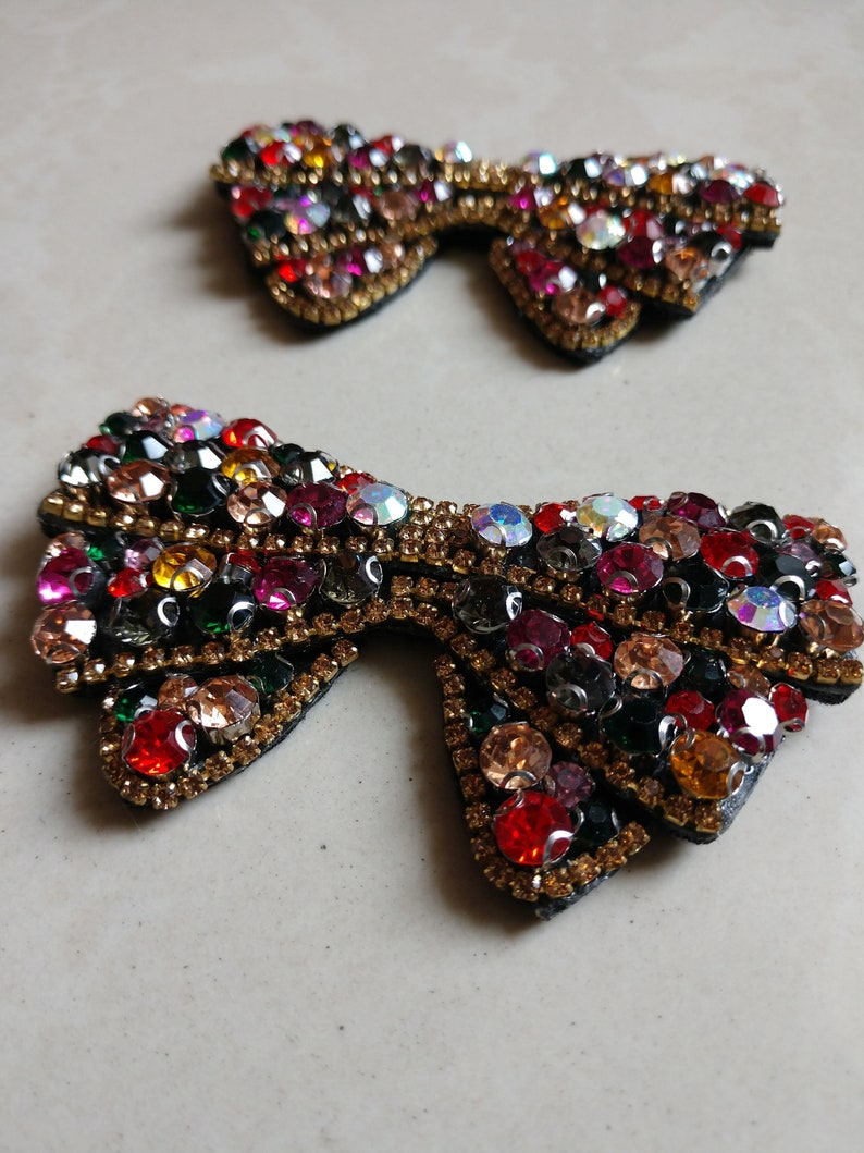 May include: Two bow-shaped embellishments, each decorated with an array of colourful rhinestones. The rhinestones are in shades of red, orange, green, and clear, set against a black base with gold trim. These decorative pieces are ideal for adding sparkle to clothing or accessories.