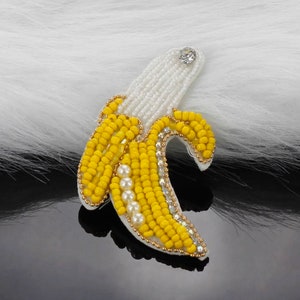Pode incluir: Um broche de banana de contas amarelas e brancas com um detalhe de strass.