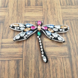 May include: A colorful dragonfly applique with a black felt body and wings covered in rhinestones and beads. The dragonfly has a green, pink, and orange body with clear, blue, and orange rhinestones.