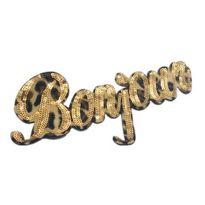 Puede incluir: Un parche decorativo con la palabra "Bonjour" en lentejuelas doradas. Las letras están cosidas sobre una tela con estampado de leopardo, creando un diseño elegante. Este parche es ideal para añadir estilo a la ropa o accesorios.