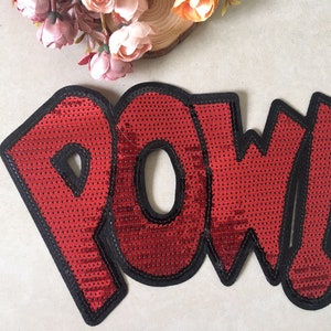 Può includere: Toppa in paillettes rosse da stirare con la parola "POW" in un font in stile fumetto.