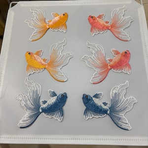 Könnte beinhalten: Sechs gestickte Goldfisch-Aufnäher in Orange, Rosa und Blau. Jeder Fisch hat ein detailliertes Design mit fließenden, durchscheinenden Flossen. Die Aufnäher sind auf einer hellgrauen Oberfläche angeordnet und zeigen die leuchtenden Farben und die aufwendigen Stickereien.